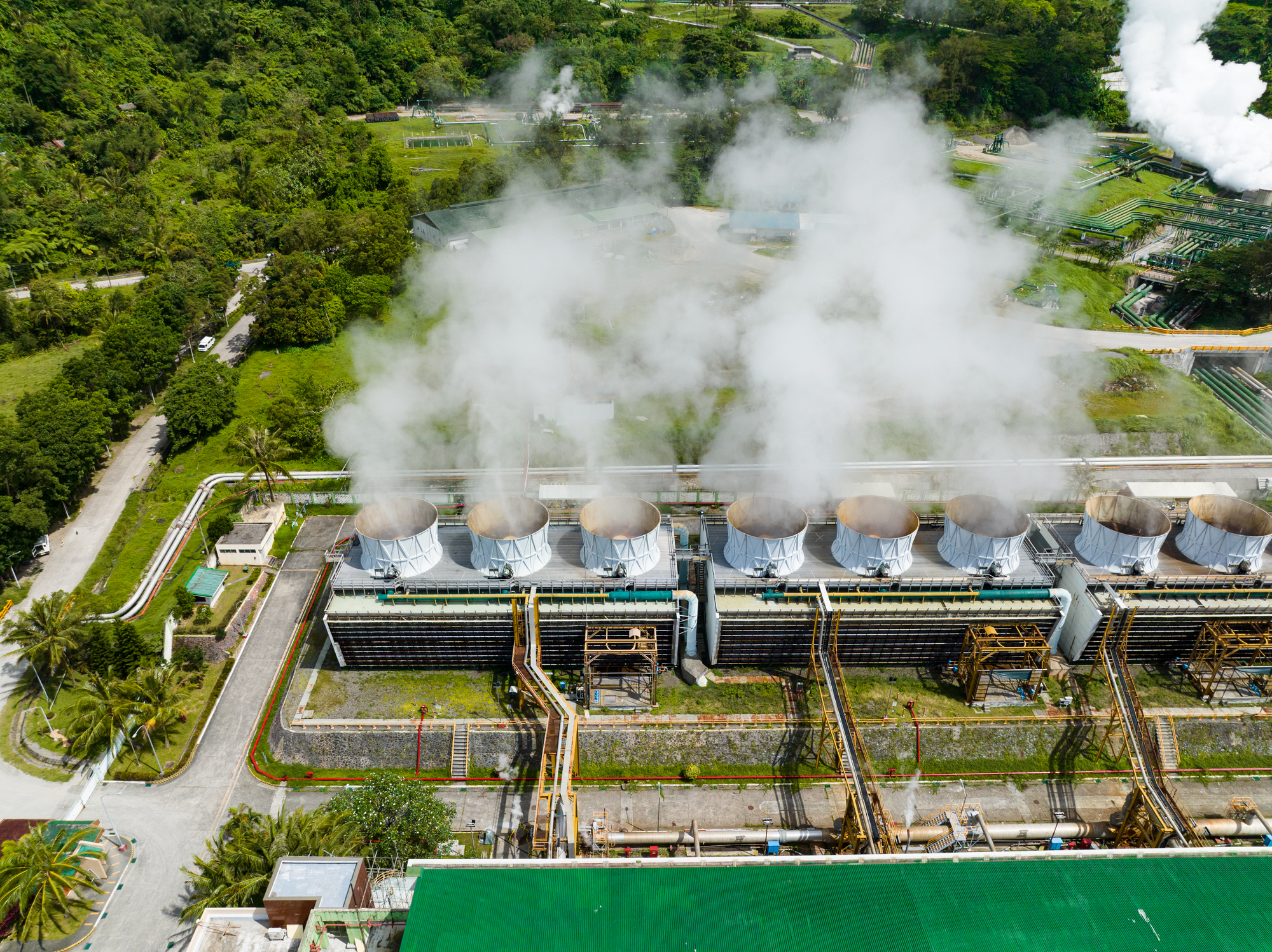 Aerial view of an industrial cooling plant with cylindrical cooling towers emitting white steam, surrounded by greenery and pipelines.