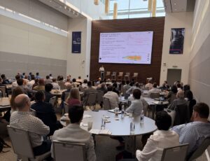 Large conference room with a speaker presenting a slide deck to an audience seated around circular tables.